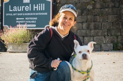 A woman in a gray hat and black sweatshirt smiles as she kneels on the ground next to a small white and brown dog.